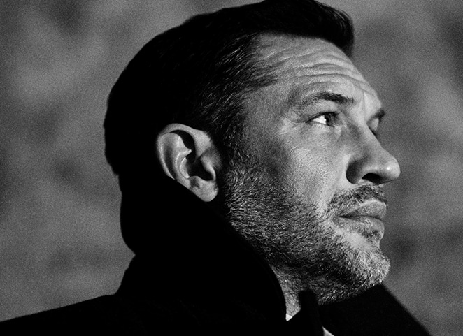 A side portrait of Tom Hardy in black and white, gazing into the distance. He wears a formal coat, with an upturned collar.
