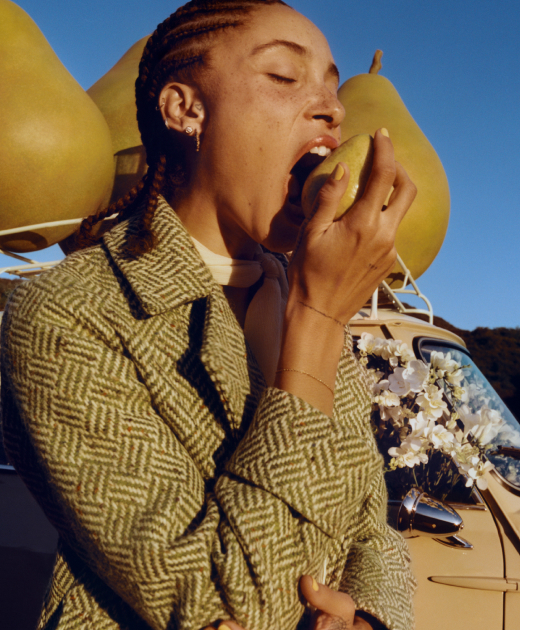 Adwoa Aboah with braided hair, biting into a pear with backdrop of three large pear props placed on top of a car roof rack.